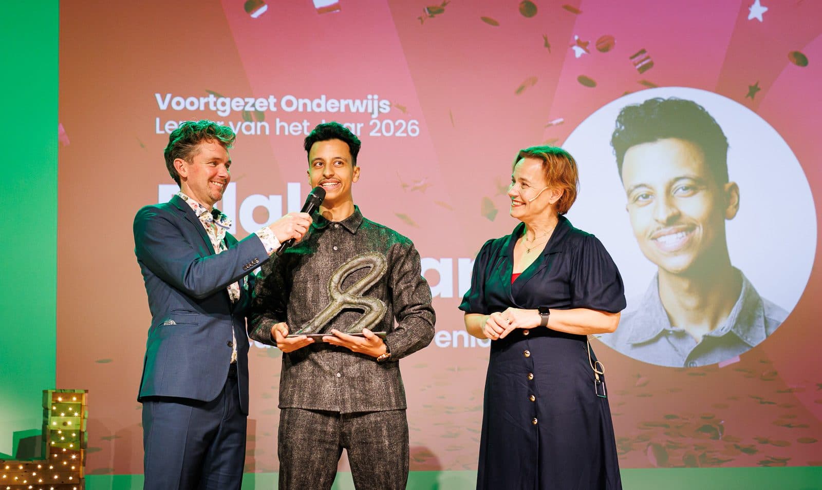 Overzicht van het podium: Bilal met trofee naast de presentatoren onder het scherm met zijn portret
