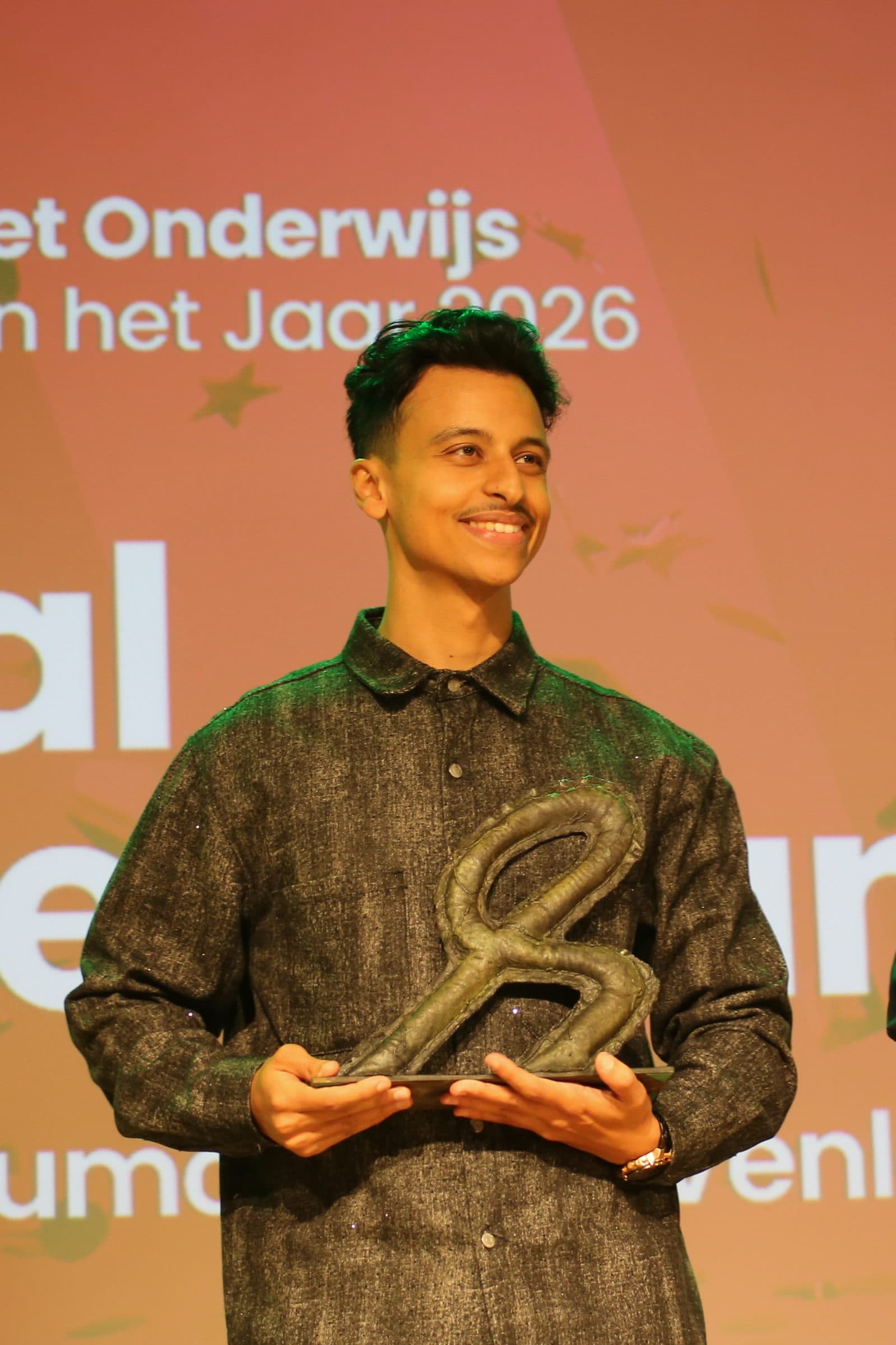 Bilal staat met zijn trofee tussen de presentatoren voor het scherm 'Voortgezet Onderwijs, Leraar van het Jaar 2026'