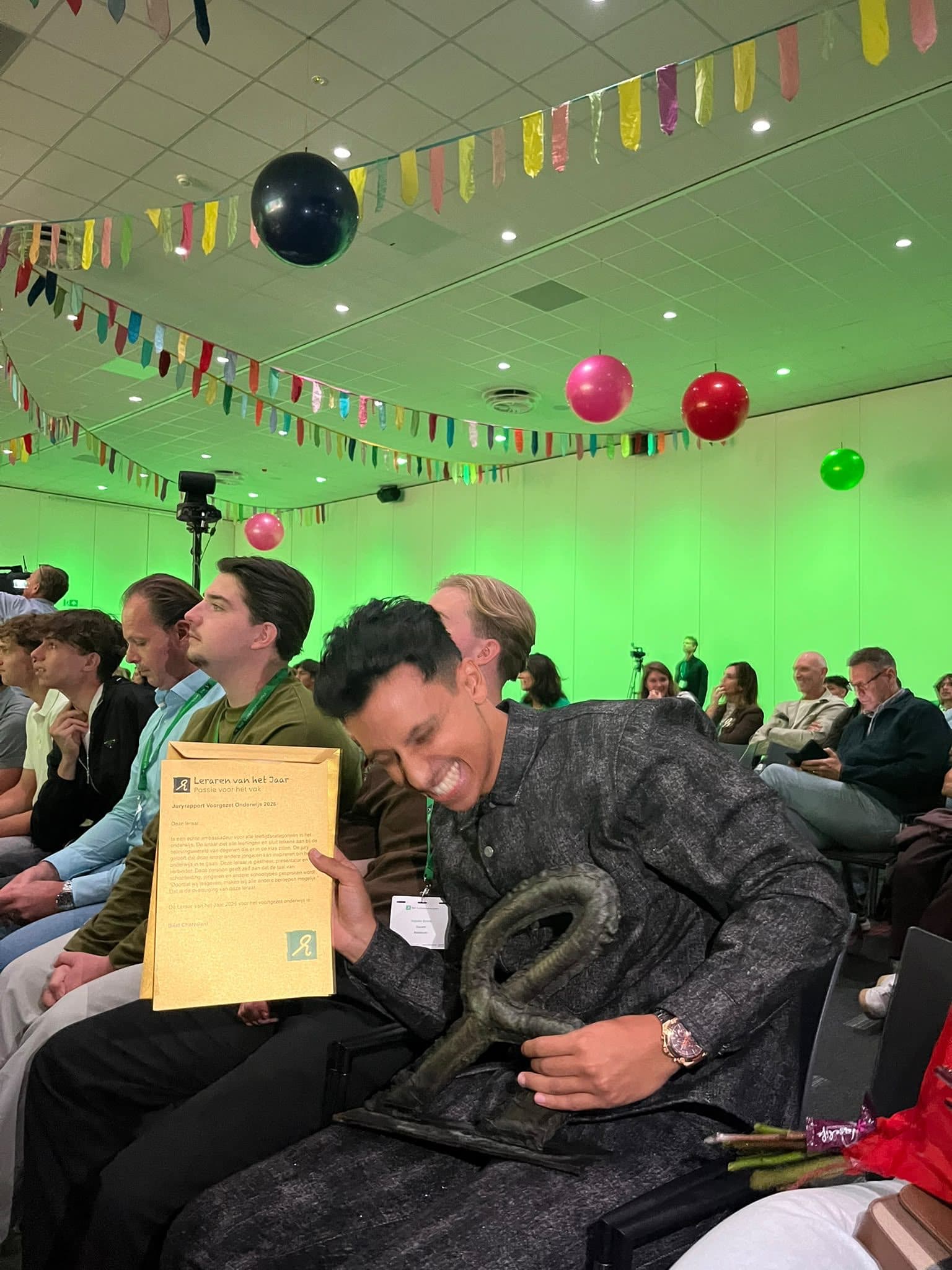Bilal in de zaal met zijn trofee en oorkonde, lachend tussen het publiek